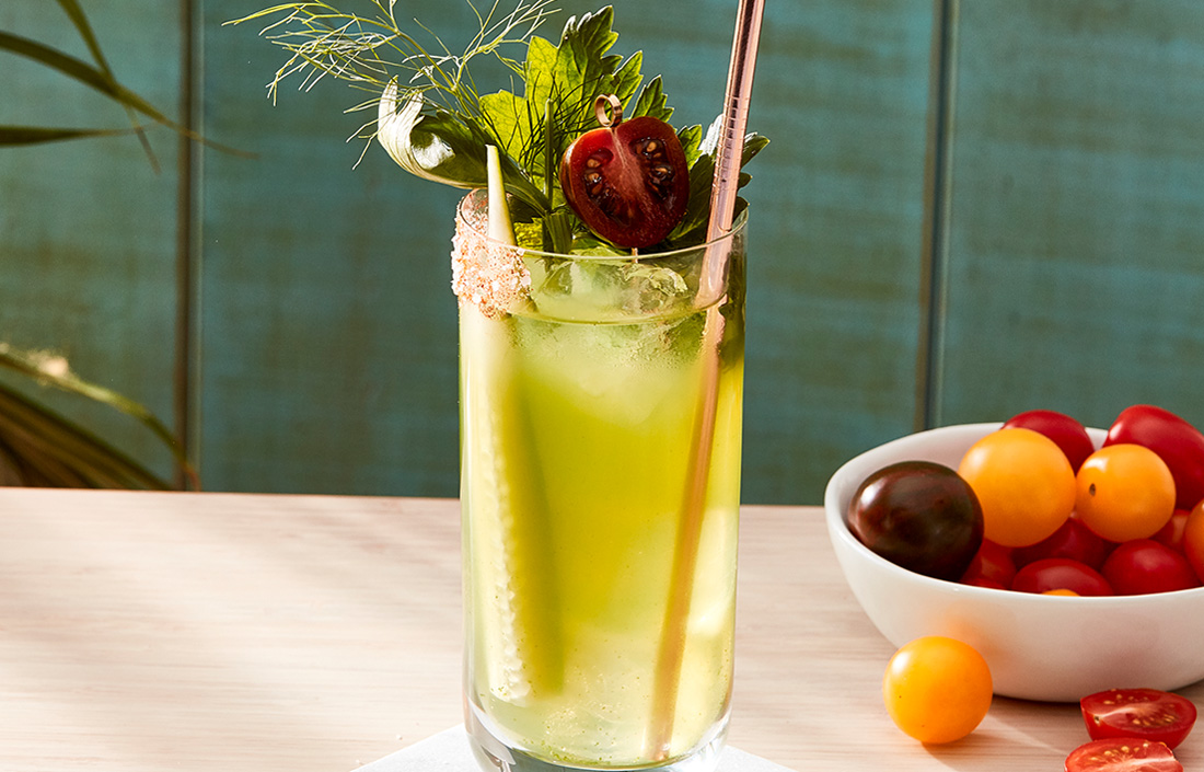 The Buchanan's green bloody marry garnished with a cucumber slice, a celery stalk, and a fresh cherry tomato next to a bowl of fruits