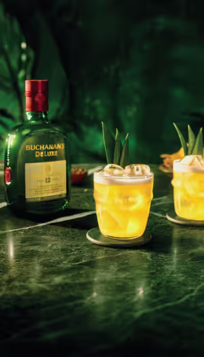 Two Buchanita cocktails on the rocks with pineapple garnish and a bottle of Buchanan's Deluxe Whiskey on a green marble counter