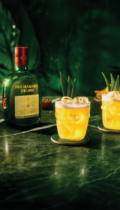 Two Buchanita cocktails on the rocks with pineapple garnish and a bottle of Buchanan's Deluxe Whiskey on a green marble counter