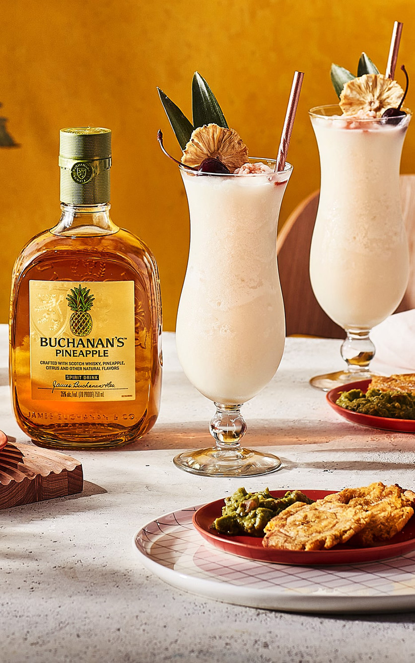 A bottle of Buchanan's Pineapple between plates of food and 2 Buchacolada drinks