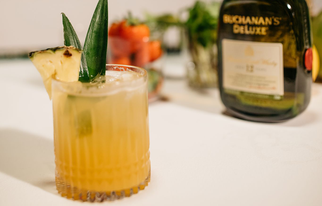 The piña crest drink garnished with a pineapple wedge in front of a Buchanan's Deluxe bottle 