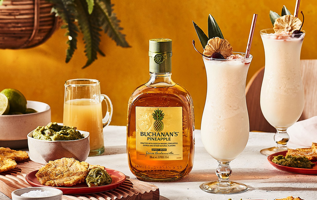 A bottle of Buchanan's Pineapple between plates of food and 2 Buchacolada drinks