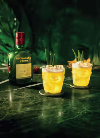 Two Buchanita cocktails on the rocks with pineapple garnish and a bottle of Buchanan's Deluxe Whiskey on a green marble counter