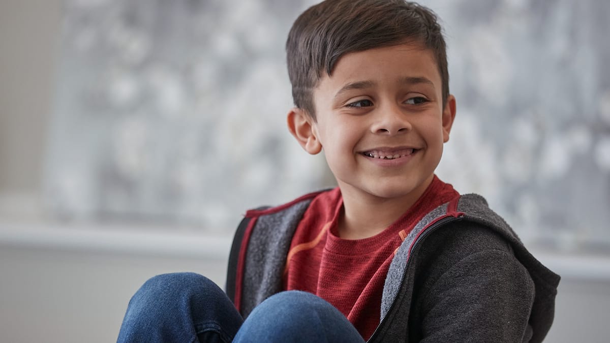 A child sitting down inside, looking to the left and smiling. The child has short brown hair, is wearing jeans, a red t-shirt and a grey hoodie. 