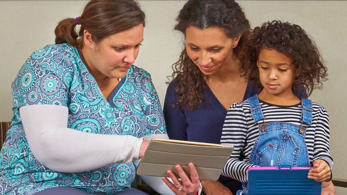 A healthcare provider showing something on a tablet device to an adult and child. 