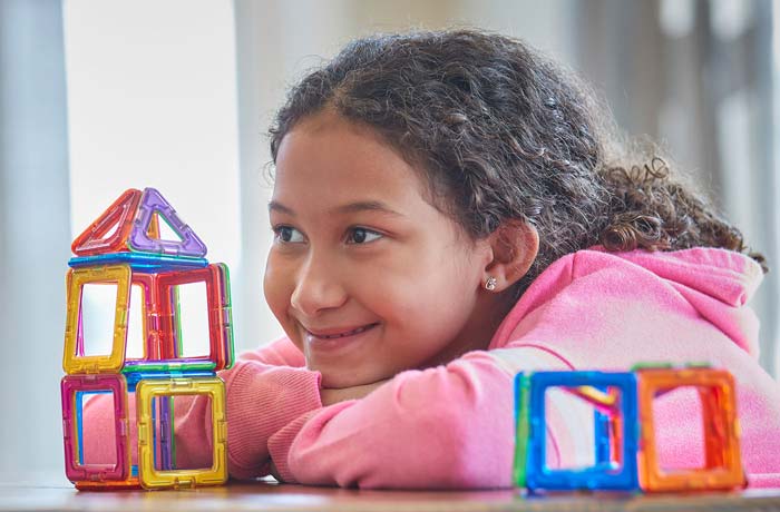 A child in a pink hoodie sitting at a table with plastic bricks on it. The child is smiling.