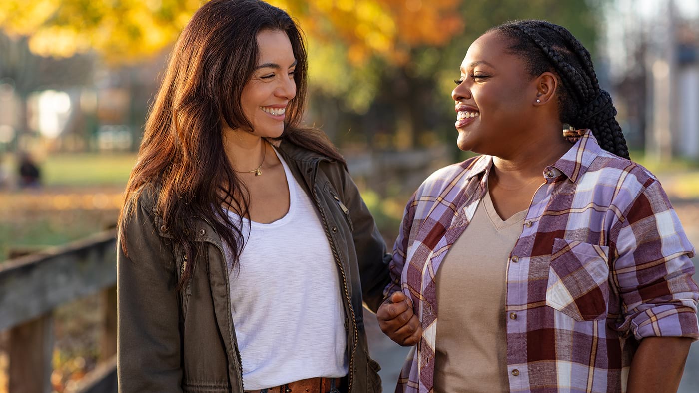 Two women on an outdoor walk. They are looking at each other and smiling. 