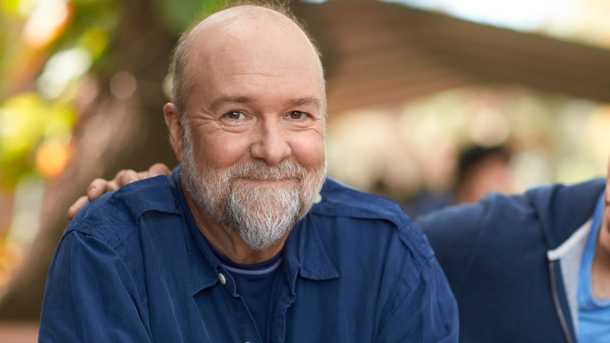 A person with a beard smiling at the camera.  He is wearing a dark blue shirt and another person has their hand on his shoulder. 