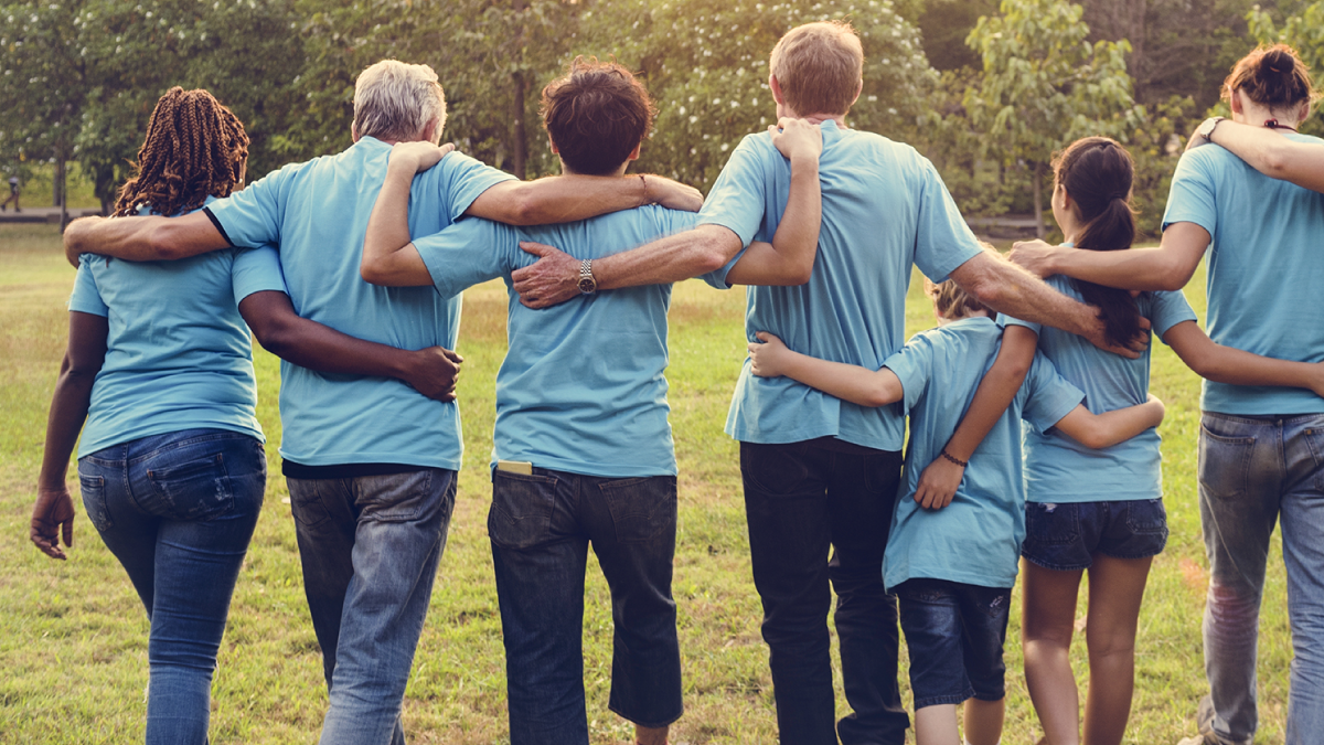 7 people of different ages standing in a line with arms interlinked behind their backs. They are outdoors and facing away from the camera. 