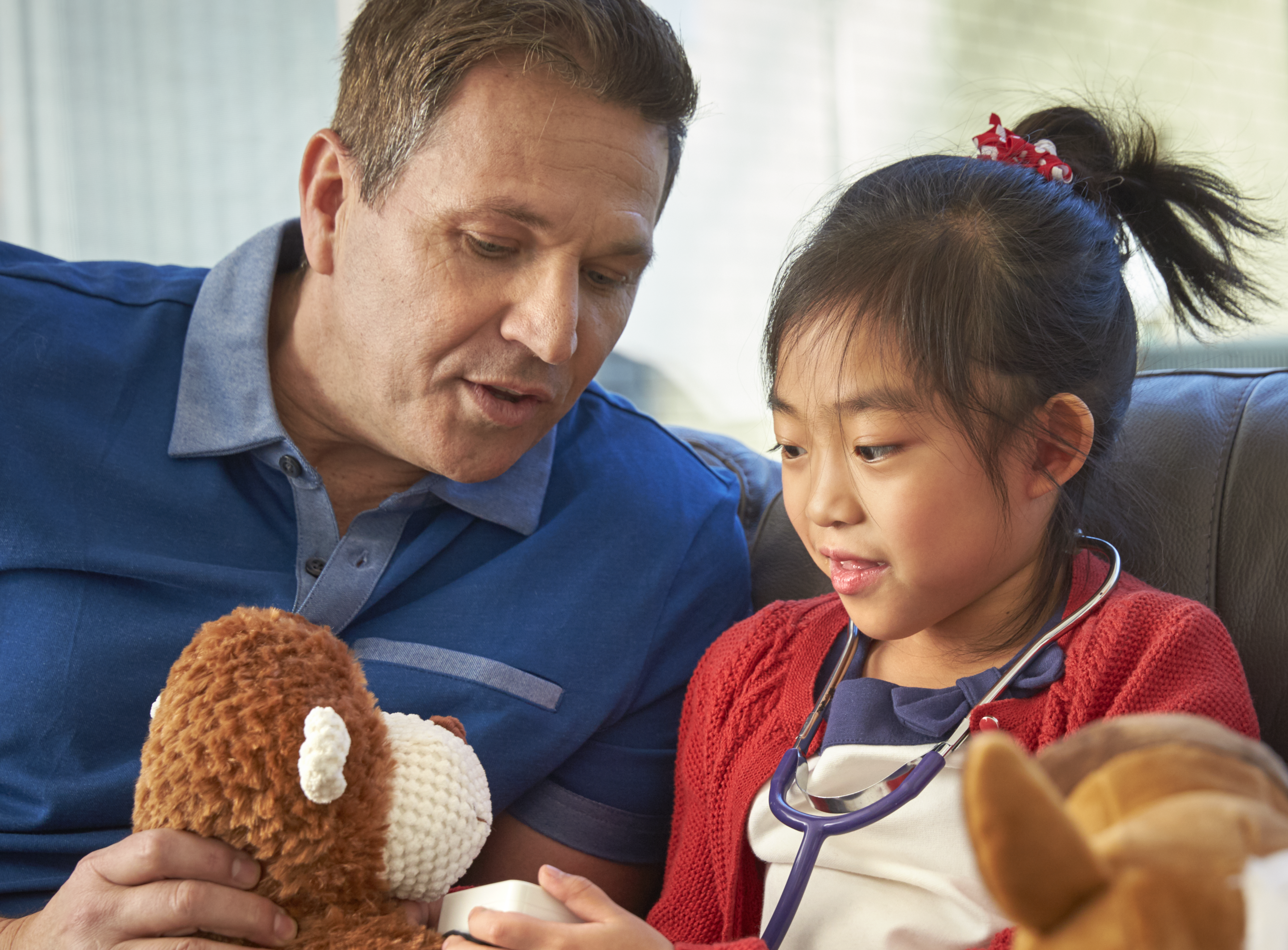 An adult holding a teddy bear and playing with a child who has a stethoscope. Both are smiling. 