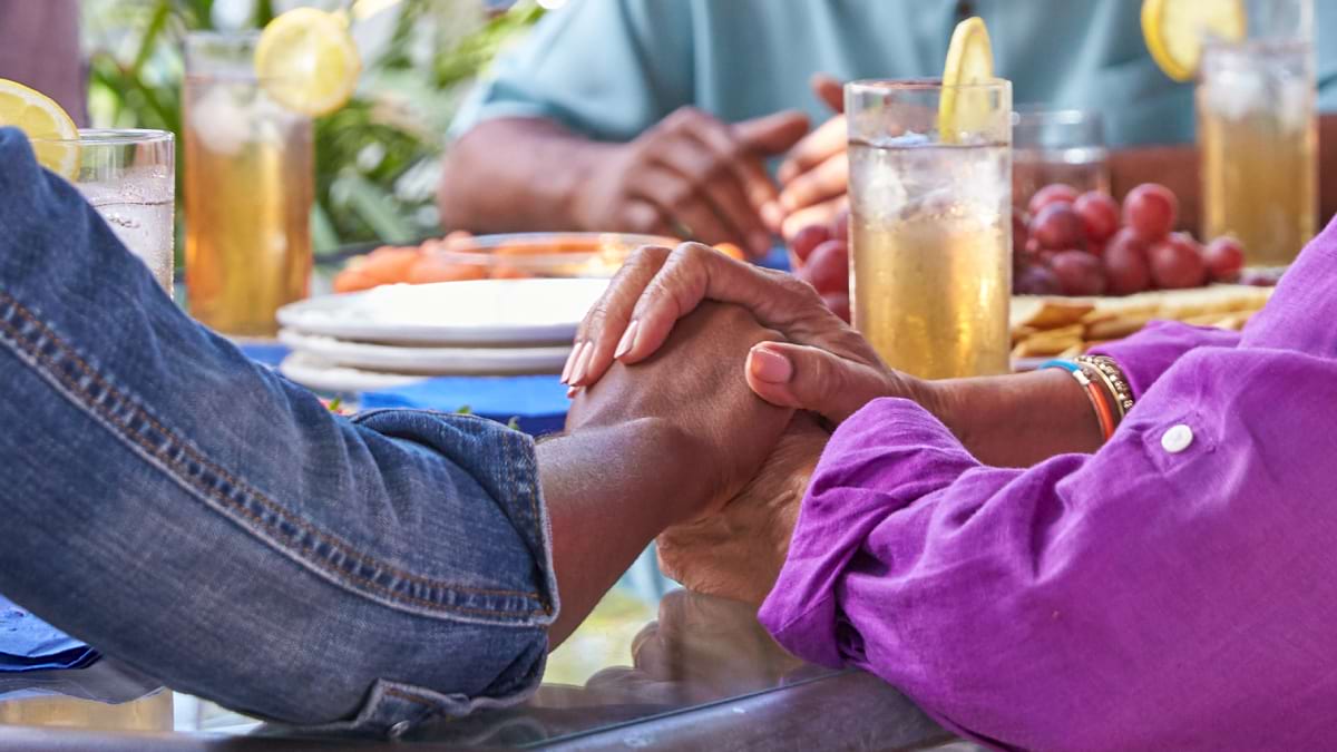 Close up of two people holding hands and supporting each other. They are sitting at a table outdoors. 
