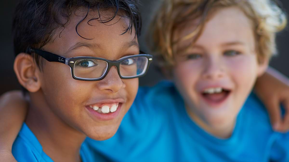 Two children linking shoulders and looking at the camera. They are both wearing blue t-shirts and smiling. 