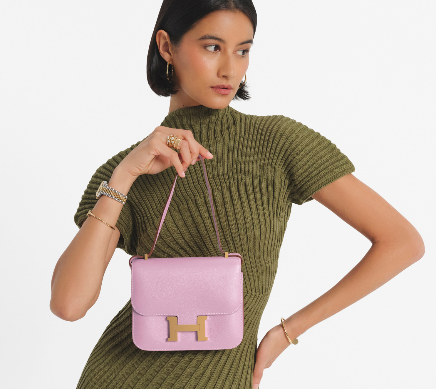 a woman with short dark hair wearing an olive green short sleeve dress and holding a light pink Hermes Constance size 18 bag with gold hardware