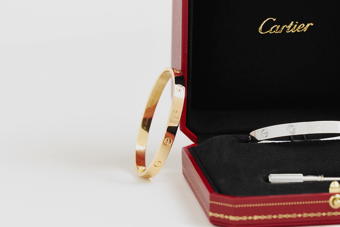 a gold Cartier Love bracelet propped against a red Cartier bracelet case with a silver Cartier Love bracelet sitting in the case