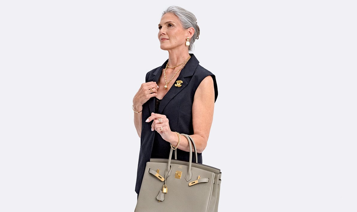 a woman wearing a black sleeveless blazer and slip dress with a gold Chanel CC brooch, a gold Tiffany Hardwear necklace and holding a grey Hermes Birkin 30 bag
