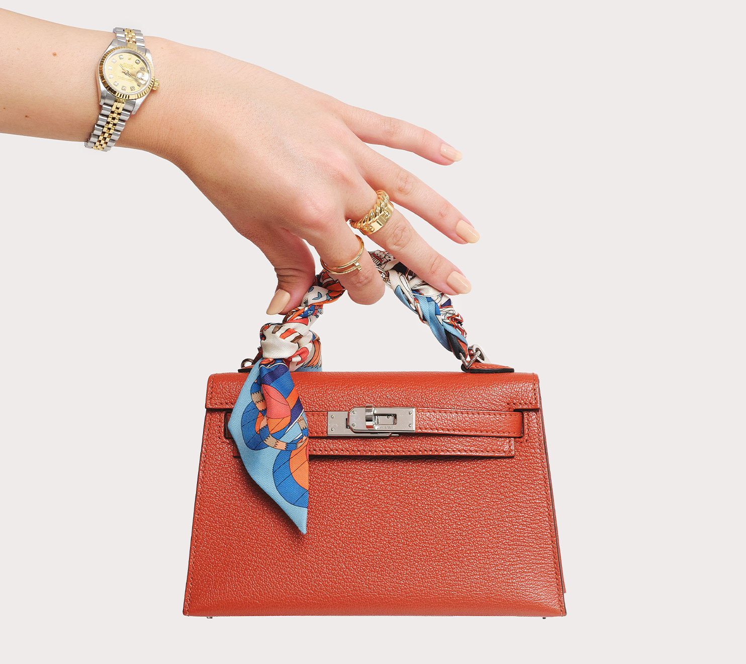 a woman's arm wearing a yellow gold and stainless steel Rolex Oyster Perpetual Watch and holding a dark orange Hermes mini kelly bag with a silk Hermes Twilly scarf wrapped around the handle