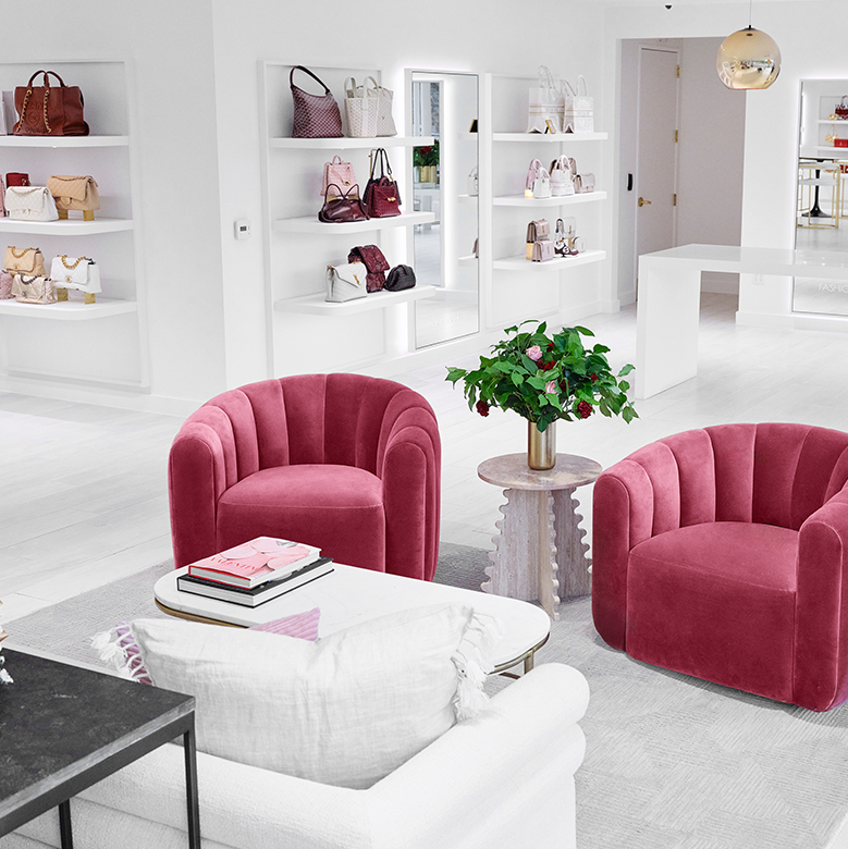 the interior of the San Diego Flagship viewing two dark pink upholstered chairs with a small side table in between and a white sofa with various designer bags displayed on the walls