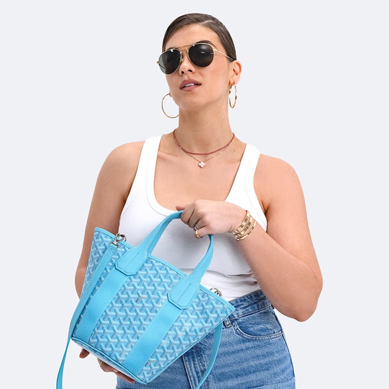 a woman wearing a white sleeveless tank top and light wash blue jeans holding a turquoise Goyard bag