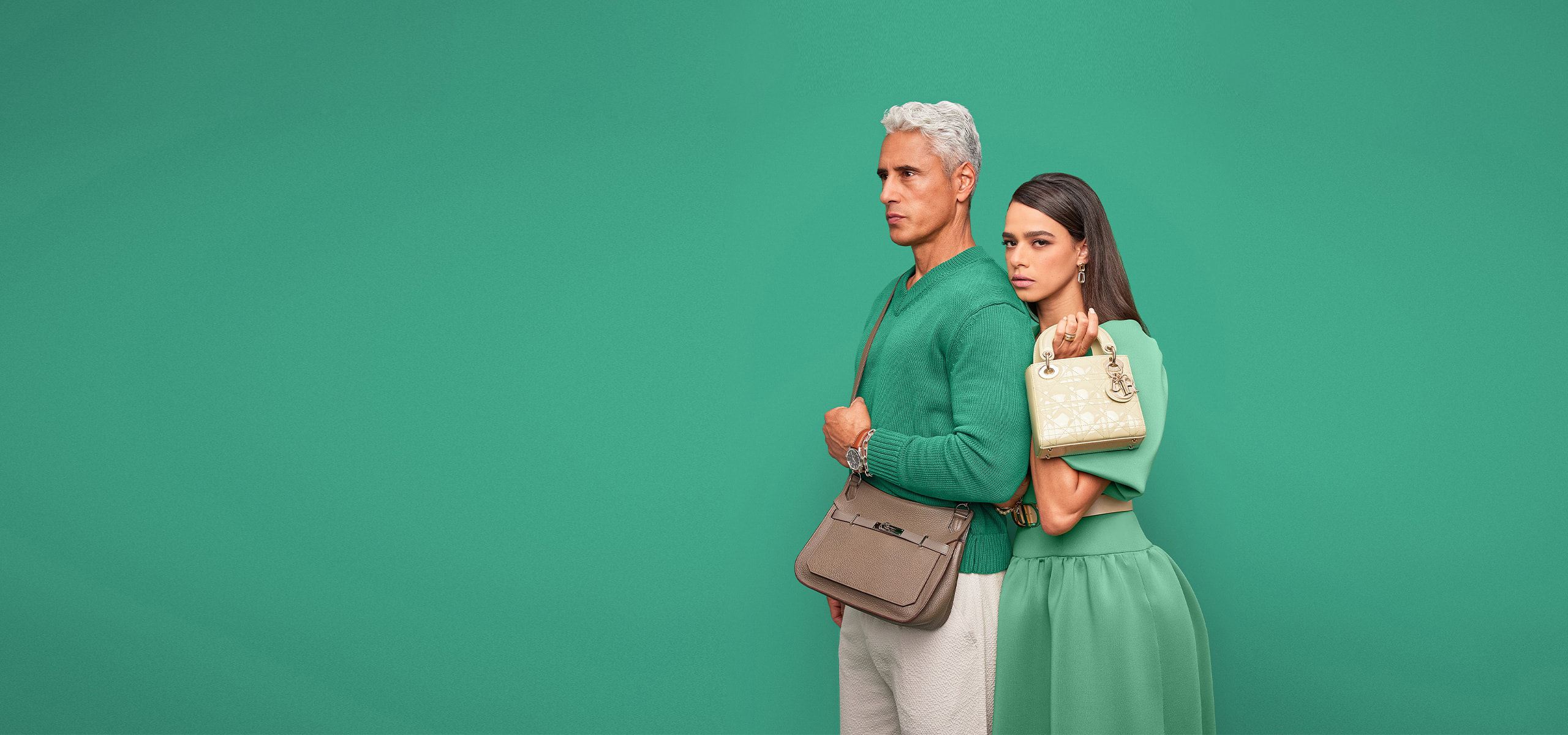 a man in a black turtleneck sweater and black pants holding a black Louis Vuitton monogram Keepall duffle bag. And a woman in a red short sleeve dress holding a white iridescent Chanel double flap bag