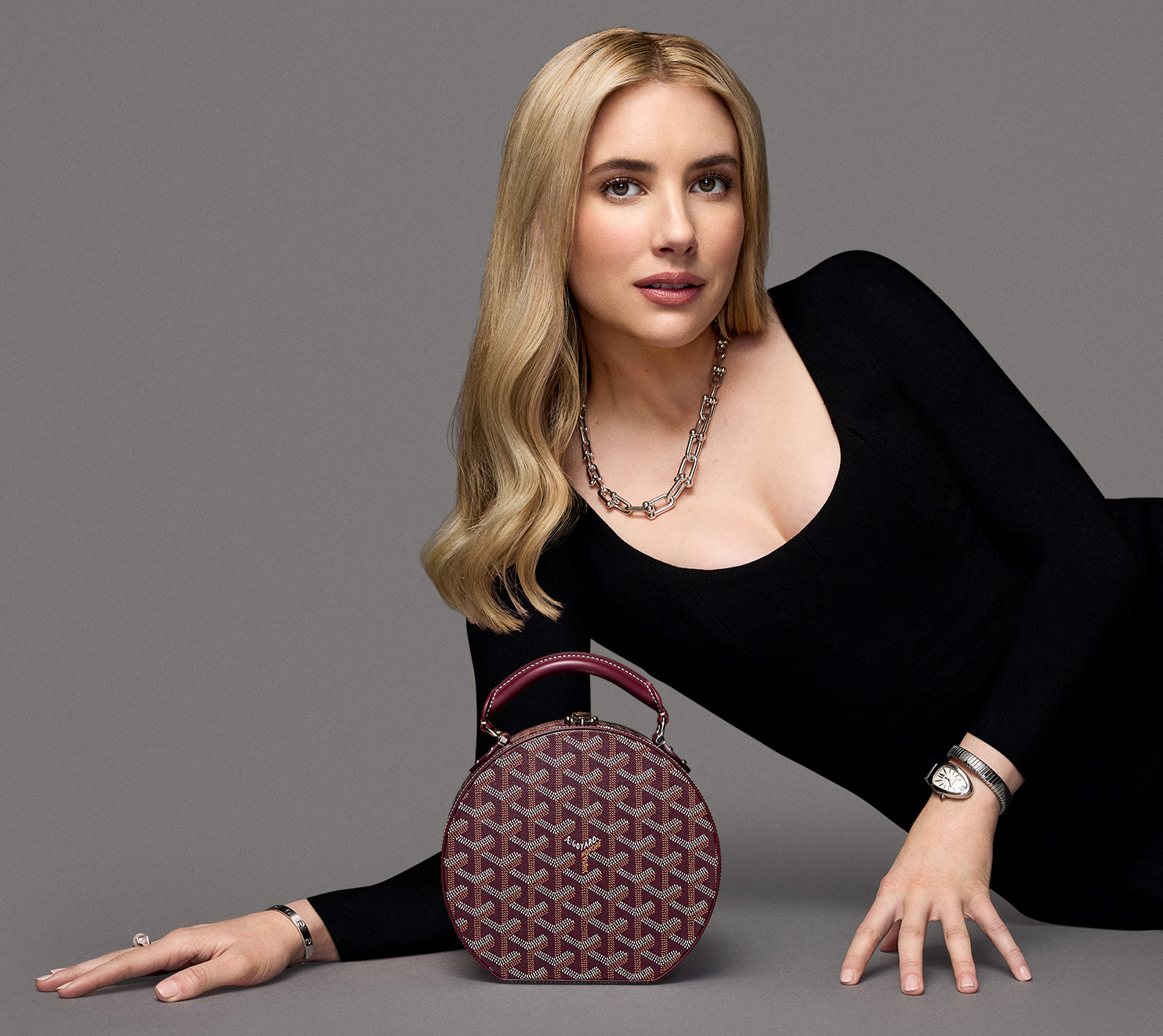 Emma Roberts wearing a long sleeve black maxi dress, a silver Bulgari Serpenti watch, a silver Tiffany Hardwear necklace, and leaning next to a burgundy Goyard Goyardine Alto Hatbox bag