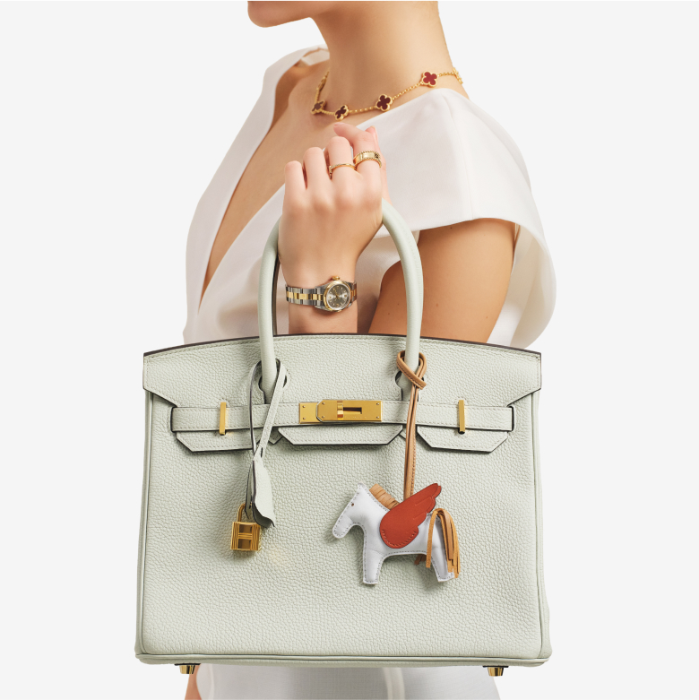 a woman wearing a white sleeveless dress holding an Hermes Birkin with an Hermes Rodeo Horse Bag Charm hanging on it