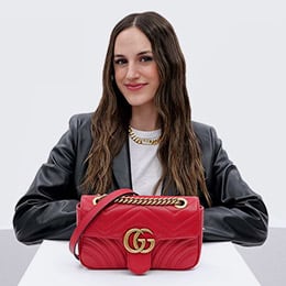 a woman wearing a black leather blazer jacket and a white t-shirt standing behind a red Gucci Marmont bag on a pedestal