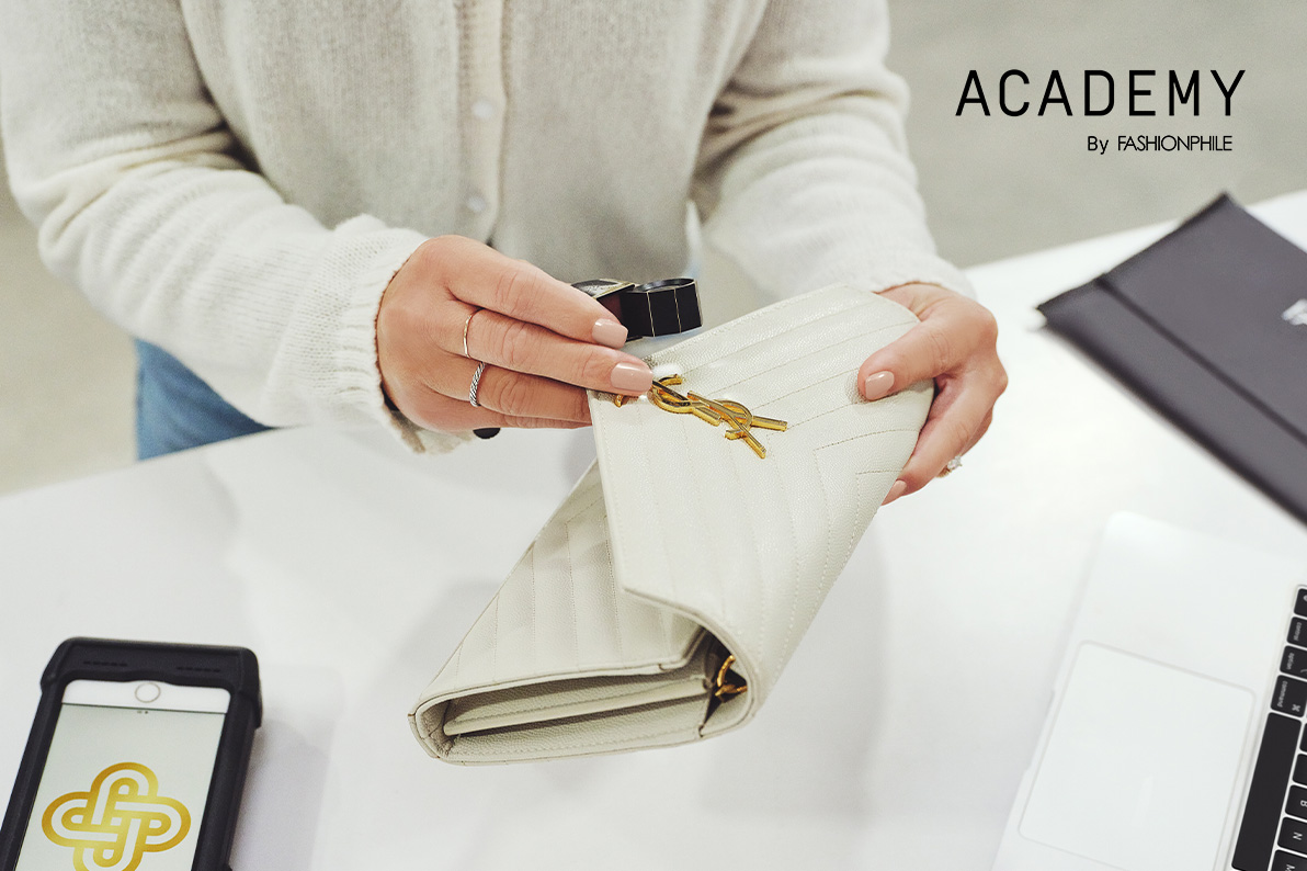 a woman wearing a white cardigan sweater and light blue jeans sitting at her desk holding and inspecting a Saint Laurent  Grain De Poudre Matelasse Chevron Monogram Chain Wallet bag in Nude Powder color