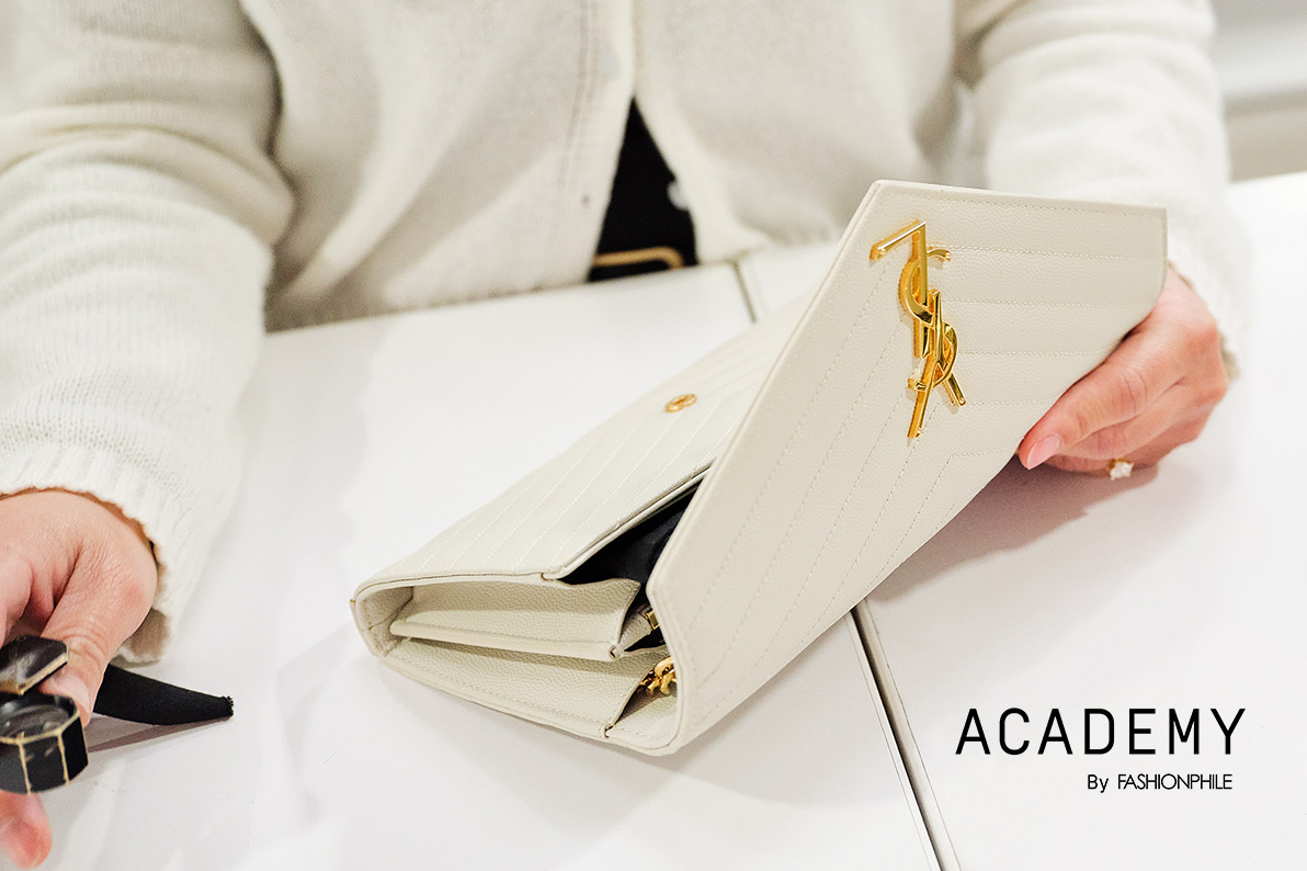 a woman wearing a white cardigan sweater and light blue jeans sitting at her desk holding and inspecting a Saint Laurent  Grain De Poudre Matelasse Chevron Monogram Chain Wallet bag in Nude Powder color