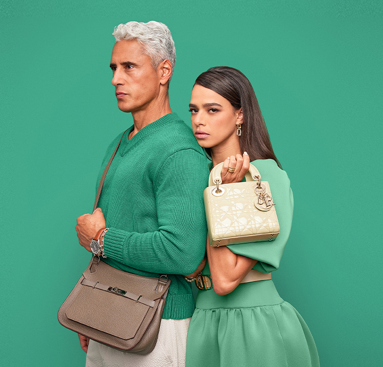 a man in a black turtleneck sweater and black pants holding a black Louis Vuitton monogram Keepall duffle bag. And a woman in a red short sleeve dress holding a white iridescent Chanel double flap bag