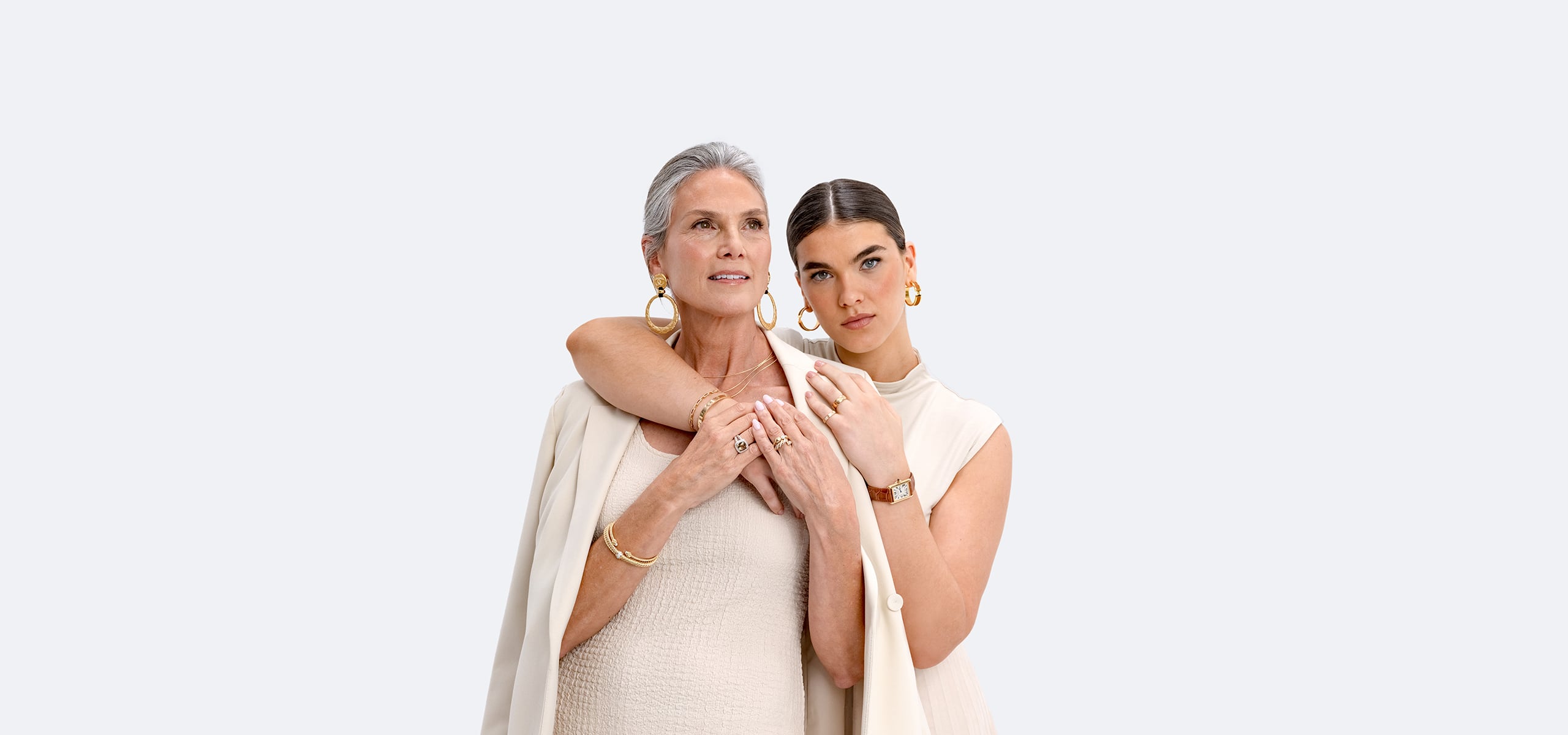 one woman wearing a beige sleeve dress with a beige blazer over her shoulders and wearing gold Chanel hoop drop earrings and another woman wearing a beige sleeveless dress, an Hermes tan strap watch, and Hermes gold hoop earrings