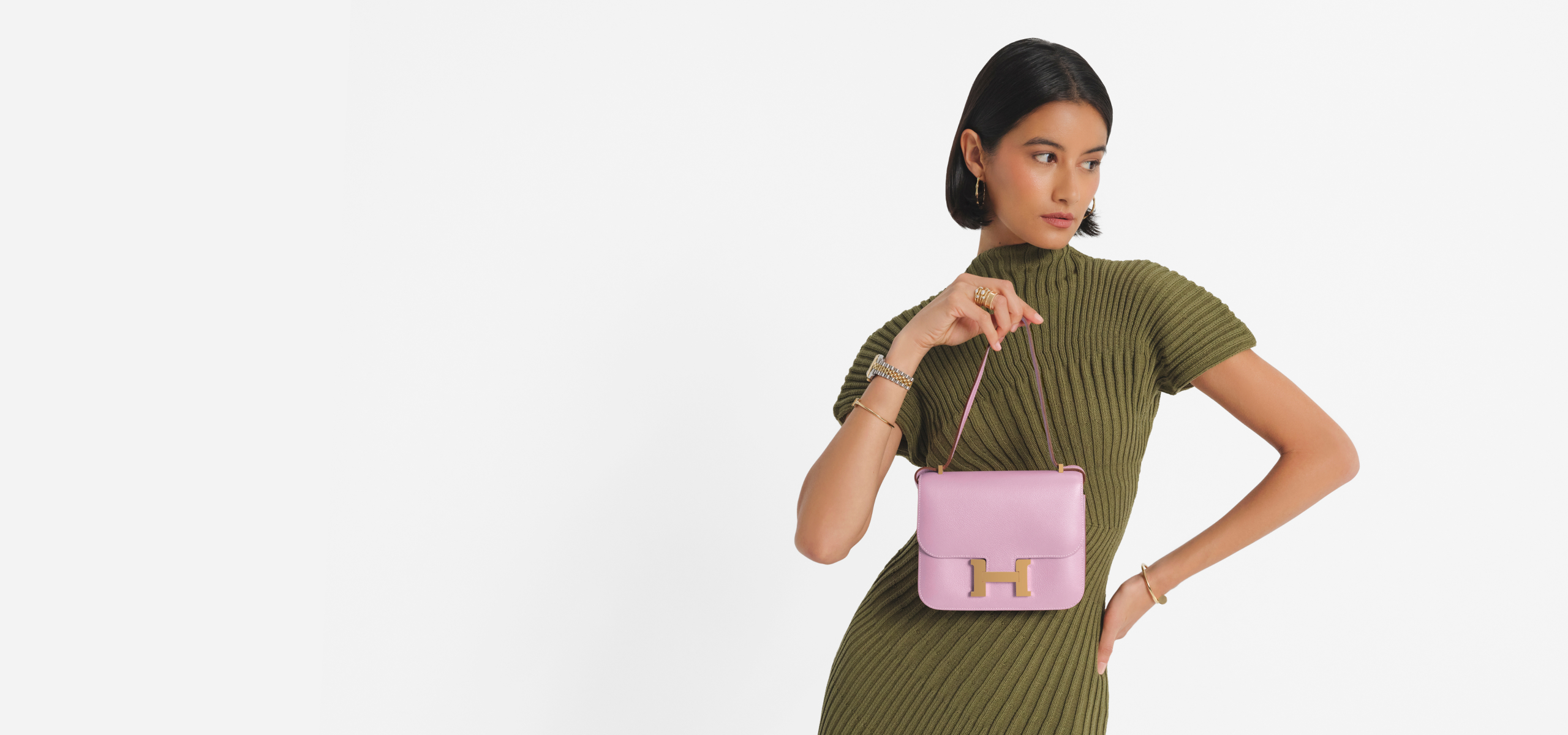 a woman with short dark hair wearing an olive green short sleeve dress and holding a light pink Hermes Constance size 18 bag with gold hardware