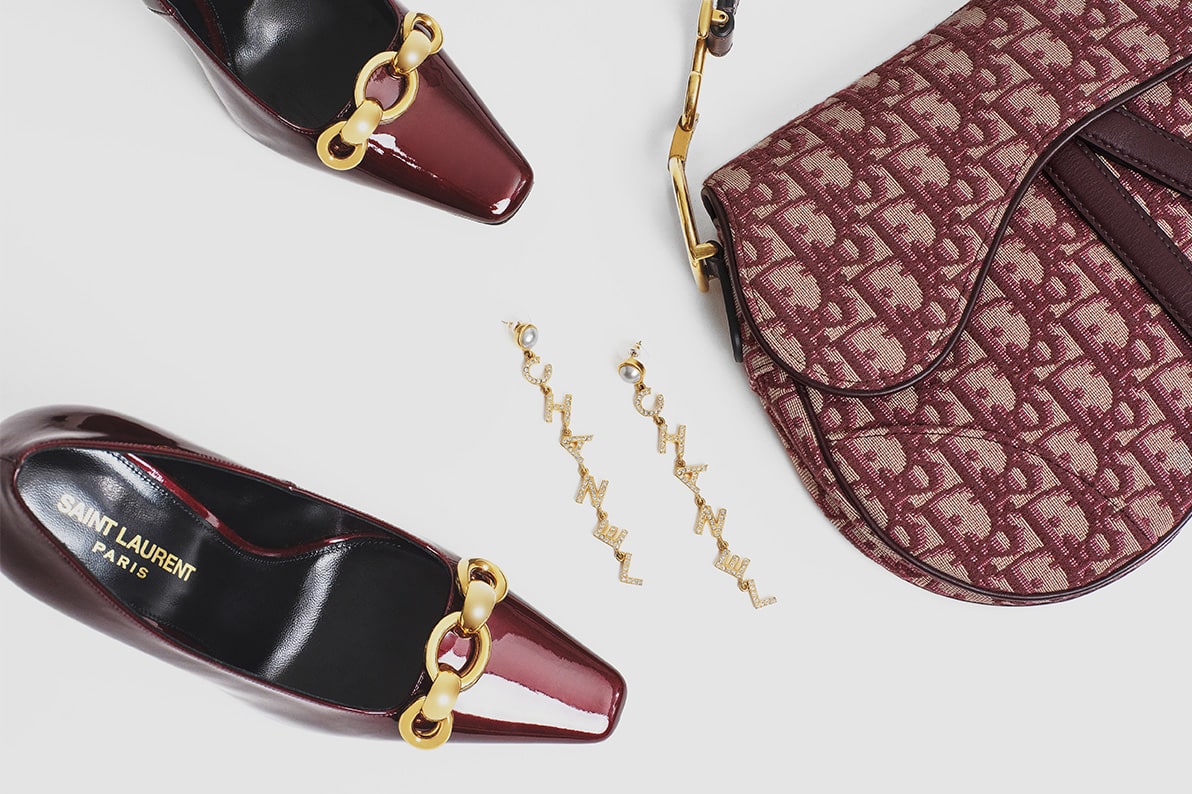 one pair of burgundy color Saint Laurent pumps with yellow gold chain hardware on the top of the foot, a pair of yellow gold Chanel crystal logo drop earrings, and one burgundy color monogram Christian Dior Oblique saddle bag with yellow gold hardware