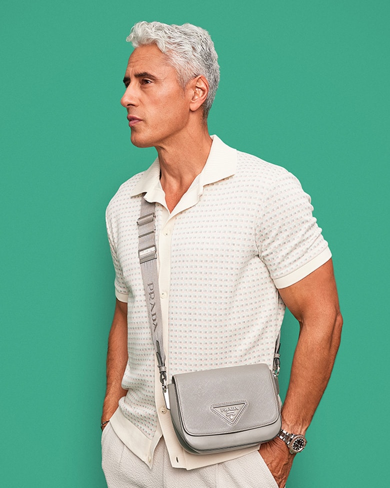 a man wearing an ecru color knit short sleeve button up shirt, ecru color slacks and a grey Prada crossbody bag