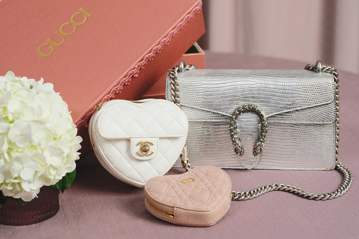 a silver Gucci Dionysus bag, a white Chanel heart shaped bag, and a beige Christian Dior heart shaped pouch sitting next to a light pink satin Gucci box and a vase of hydrangea flowers