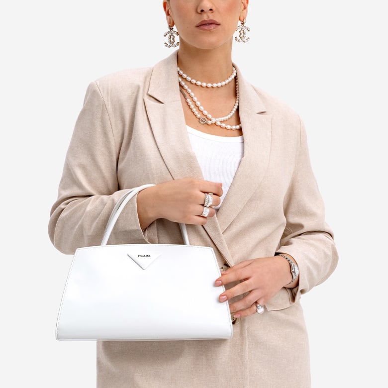 a woman in a beige blazer and Chanel drop earrings holding a white Prada Spazzolato Architectural Line Horizontal Bag 