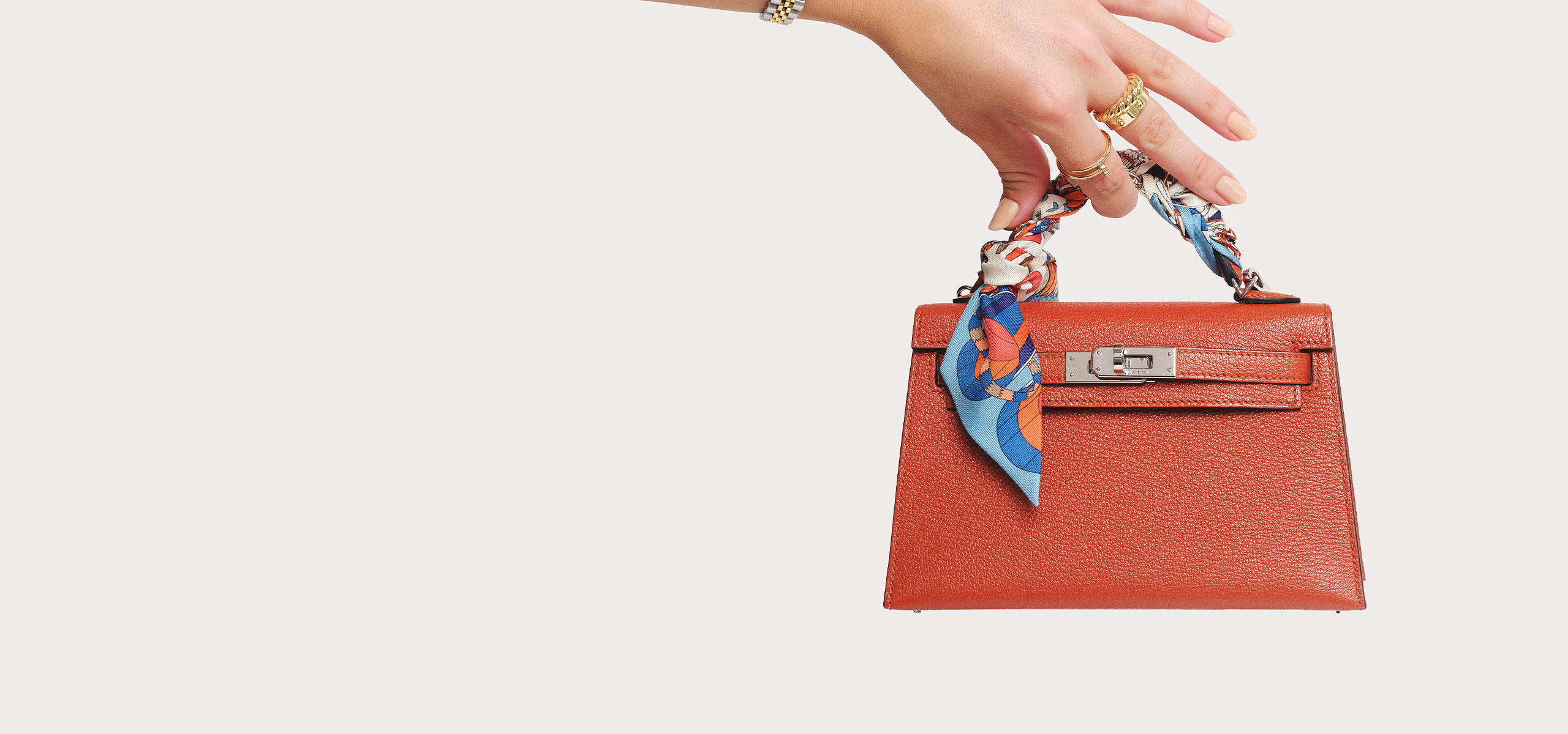 a woman's arm wearing a yellow gold and stainless steel Rolex Oyster Perpetual Watch and holding a dark orange Hermes mini kelly bag with a silk Hermes Twilly scarf wrapped around the handle