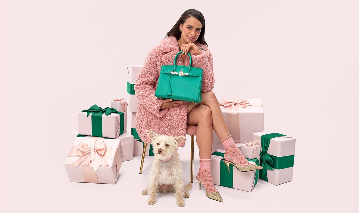a woman sitting on a chair wearing a pink shearling coat, pink monogram Gucci socks, gold crystal embellished Prada slingback pumps and holding a green Hermes Birkin 30 bag. Pink gift boxes of various sizes displayed around her.