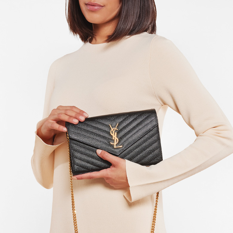 a woman in a beige long sleeve dress holding a black SAINT LAURENT Matelasse Chevron Monogram Envelope Chain Wallet 