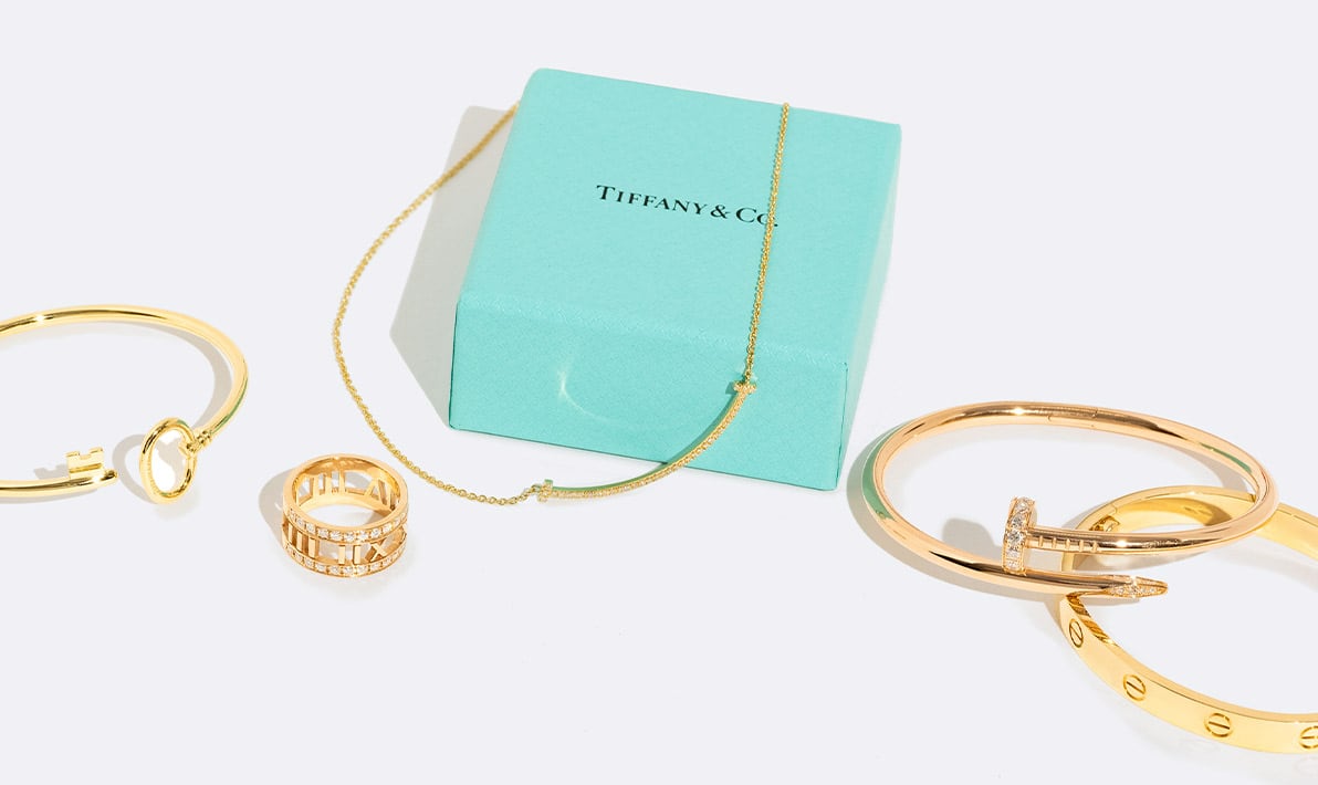 one Tiffany Gold Keys Wire Bracelet, one Tiffany gold Atlas ring, one Tiffany Gold Diamond Small T Smile Pendant Necklace laying on a Tiffany blue box, one Cartier Yellow Gold Diamond Juste Un Clou Bracelet, and one Cartier Yellow Gold Love Bracelet