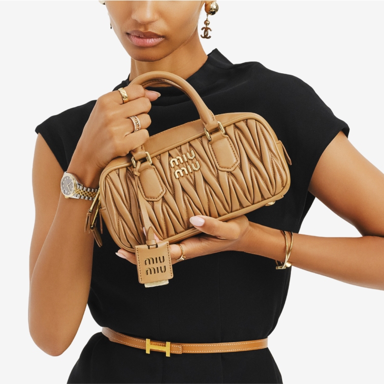 a woman wearing a black short sleeve dress with a Hermes tan H buckle skinny belt and holding a Miu Miu Nappa Matelasse Large Arcadie Top Handle Bag Caramel