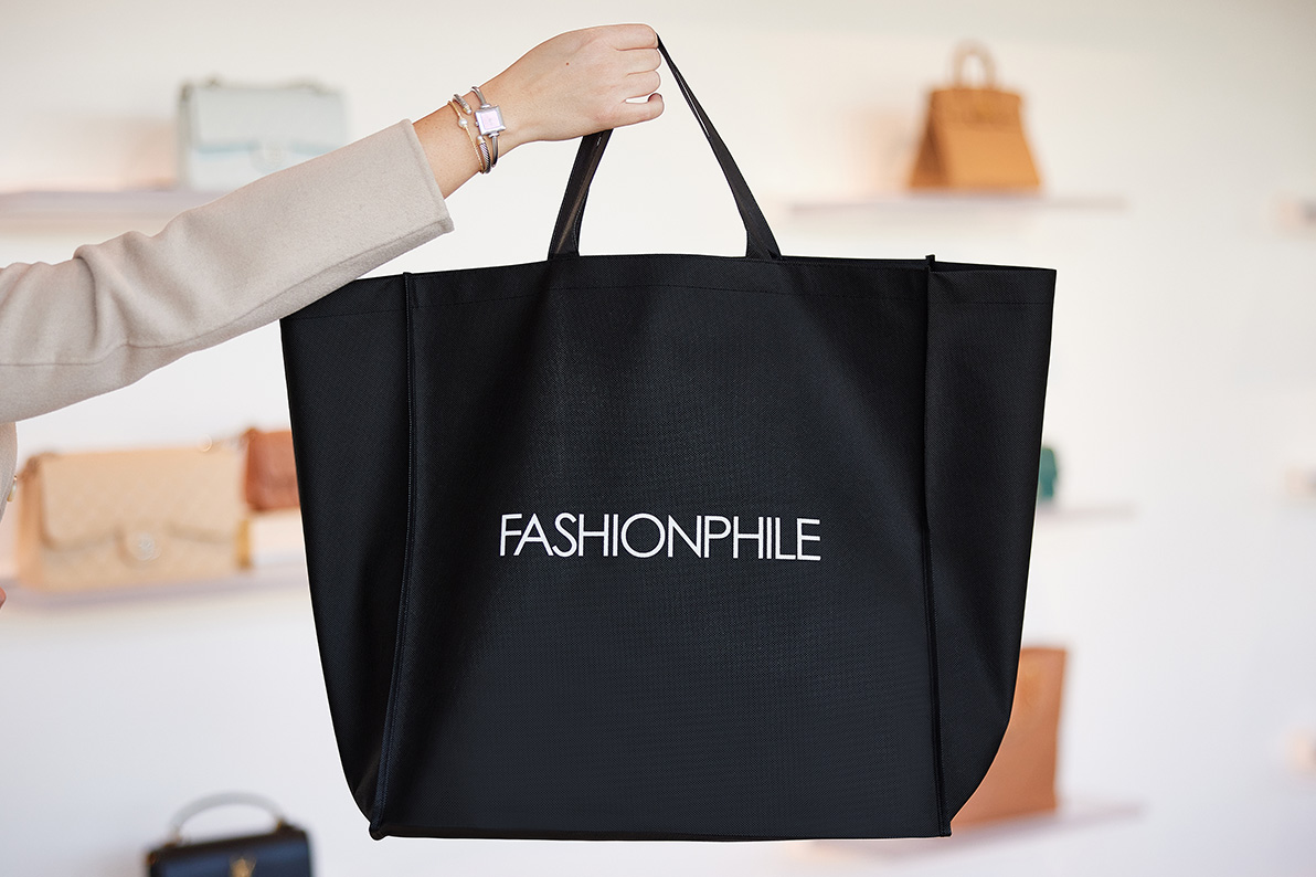 A woman in a taupe jacket holding our black FASHIONPHILE branded shopping bag in front of our bag wall at our San Diego Flagship