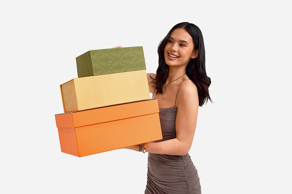 A woman wearing a grey sleeveless dress and holding an orange Hermes box, a yellow Fendi box, and a Gucci green box stacked on one another.