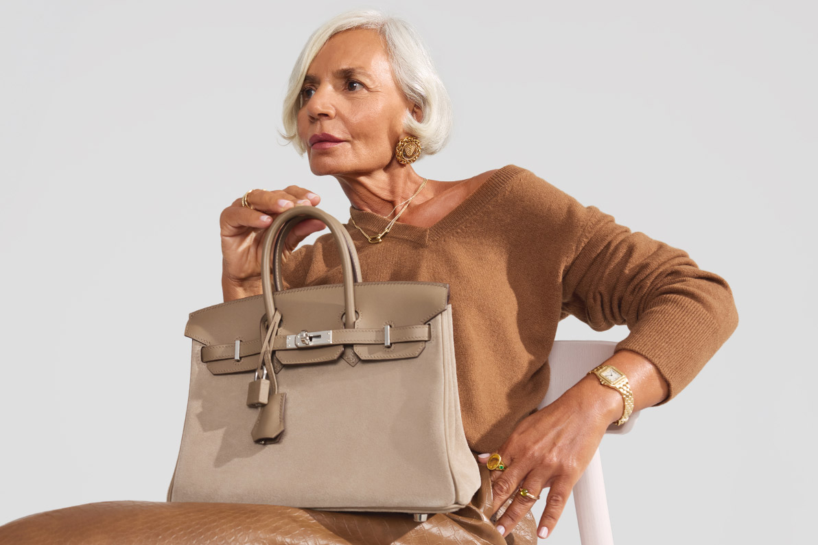 Grece Ghanem wearing a tan v-neck sweater sitting on a light pink wooden chair and holding an Hermes Veau Doblis Swift Grizzly Birkin 25 in the Etoupe color sitting on her lap