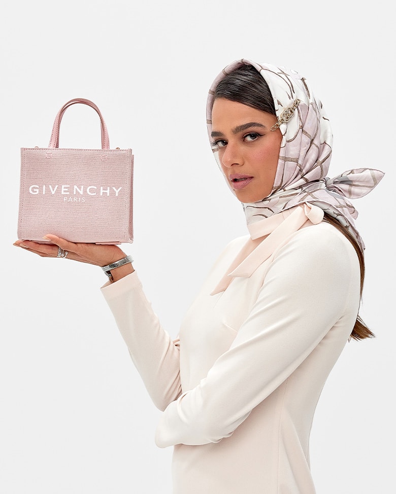 a woman in a light pink long-sleeve dress and a light pink and white patterned Chanel silk scarf wrapped around her hair holding a light pink mini Givenchy logo tote bag