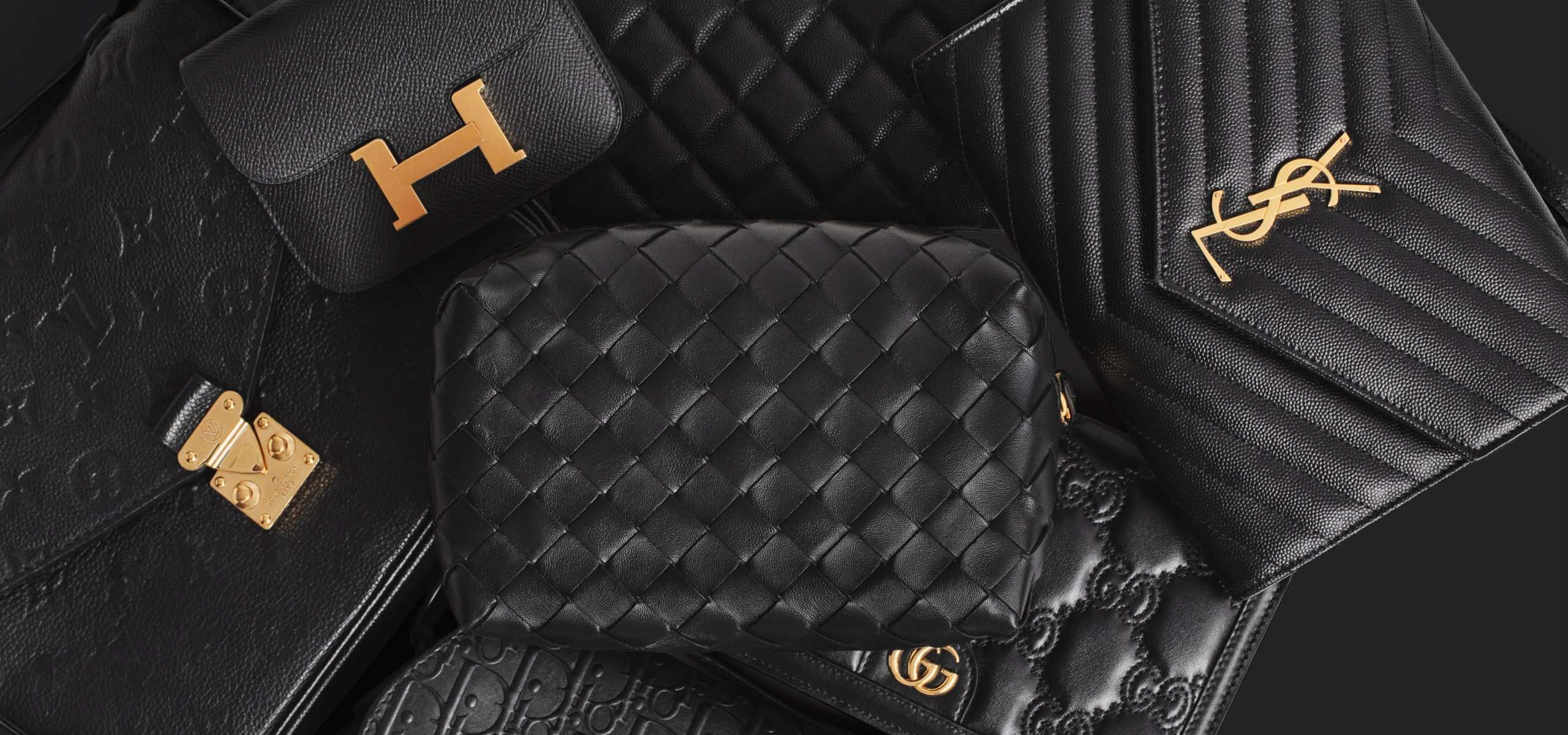 a group of black designer bags laid down - one black Louis Vuitton Pochette Metis bag, one black Hermes Constance compact wallet, one black Bottega Veneta pouch and one black Saint Laurent Chevron Quilted Envelope bag