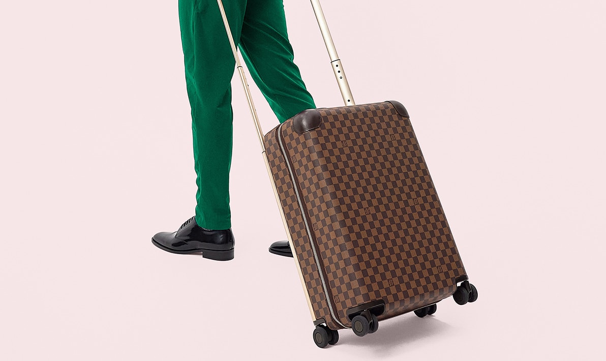 a man in a green suit and black Saint Laurent oxford shoes pulling a brown Lous Vuitton Damier Ebene pattern Horizon wheeled suitcase