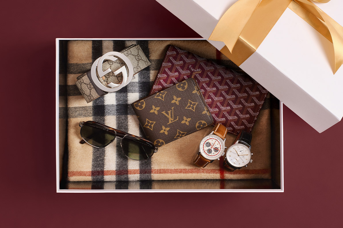 a white box with gold ribbon wrapped around the lid sitting on the corner of the box and inside is various men's designer accessories