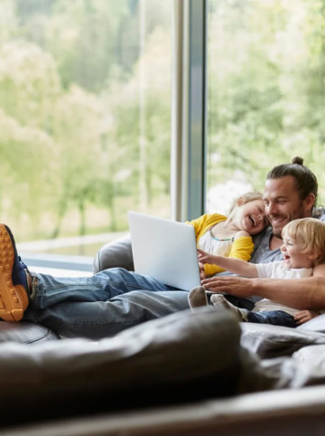 Ein Vater sitzt mit zwei Kleinkindern im Arm und ausgestreckten Beinen auf einem Sofa und schaut auf ein Laptop.