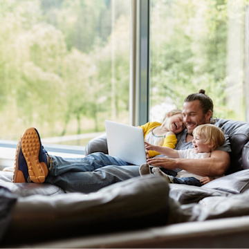 Ein Vater sitzt mit zwei Kleinkindern im Arm und ausgestreckten Beinen auf einem Sofa und schaut auf ein Laptop.