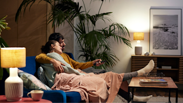 Ein junges Paar sitzt im Wohnzimmer Arm in Arm auf dem Sofa und schaut etwas auf dem Fernseher.
